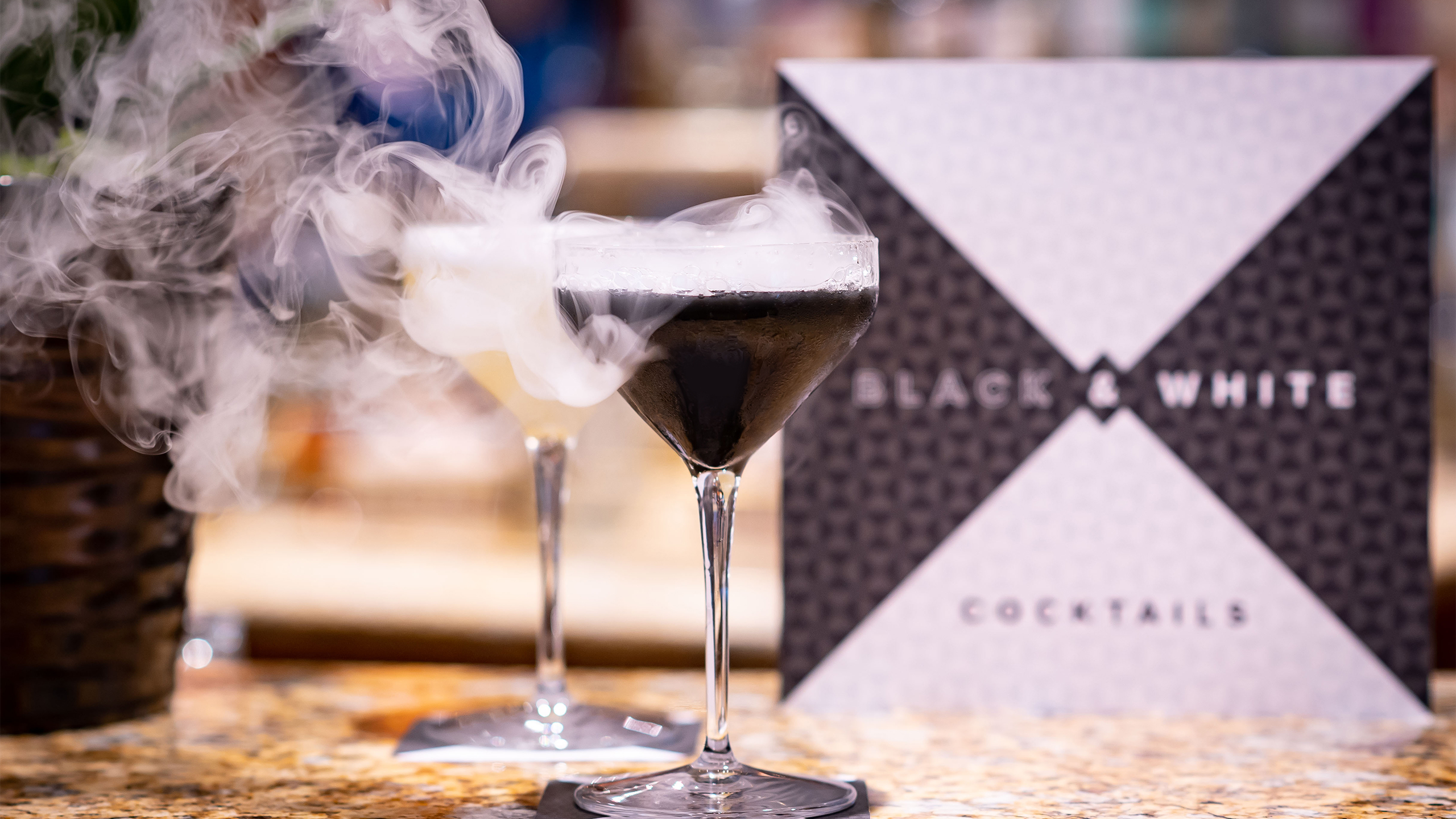 Black and White Cocktails menu standing behind two smoking cocktails on a bar top